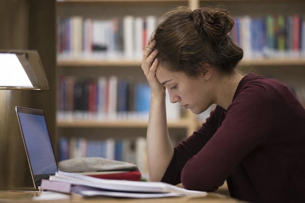 A student studying at a desk with head in hand, looking down at a laptop