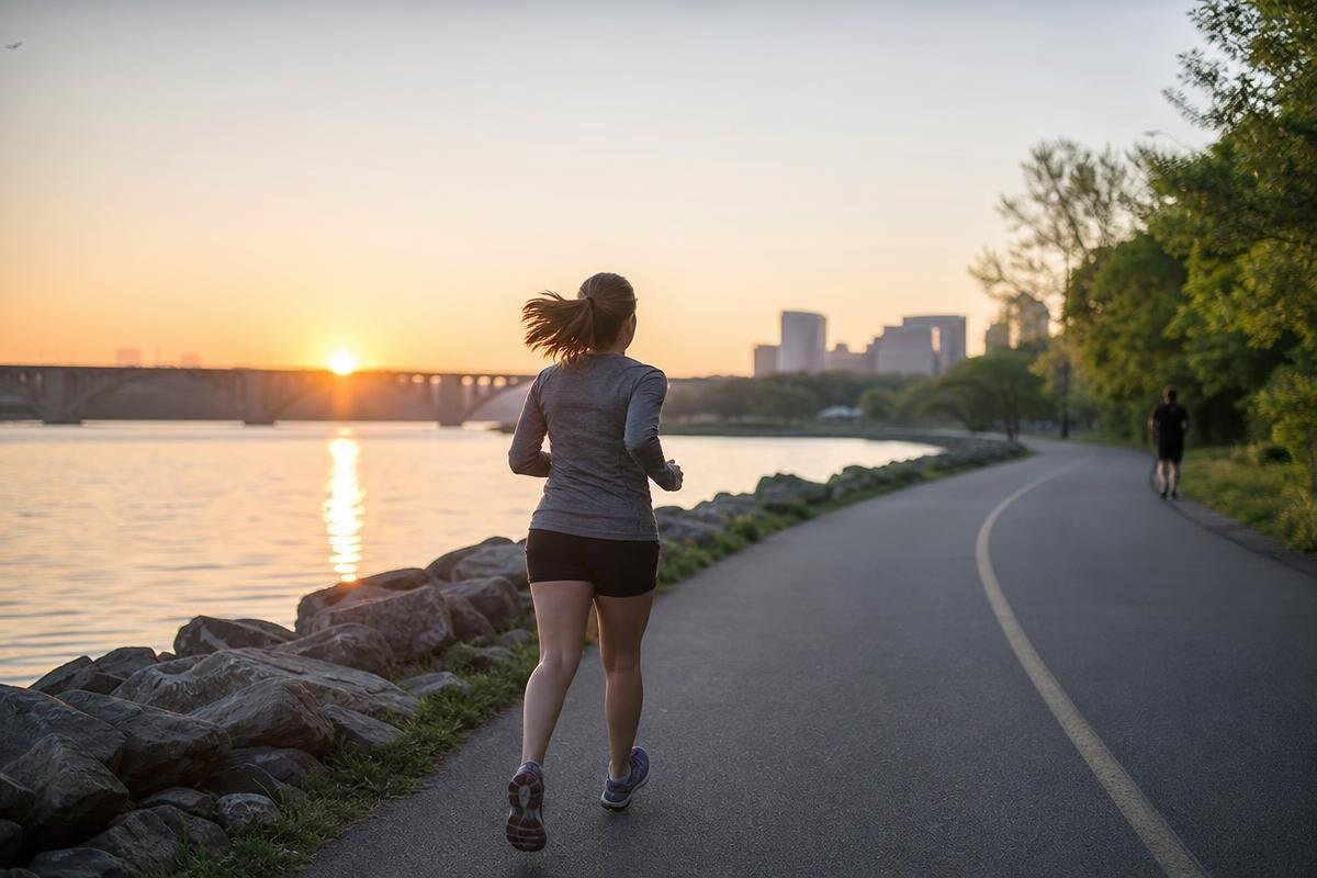 Woman running along a path by a river at sunset with a city skyline in the distance